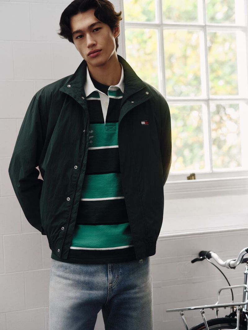 A stylish young man poses indoors, wearing a green jacket and striped shirt, standing next to a bicycle in a bright, natural light setting.