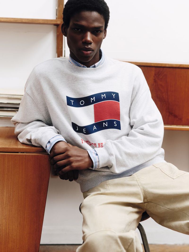 A Black male model poses indoors, wearing a Tommy Jeans sweatshirt and khaki pants, in a portrait shot with soft natural lighting.