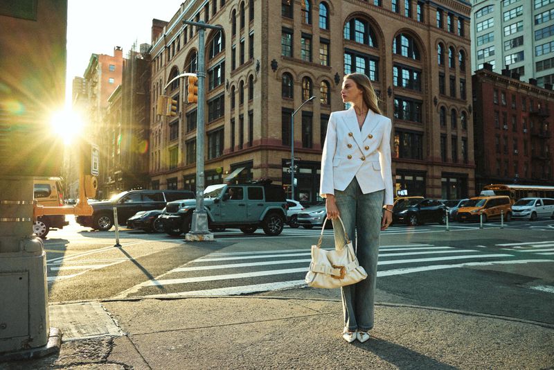 Fashion shot of a woman in a white blazer and denim pants walking across a crosswalk with city buildings and traffic in the background.