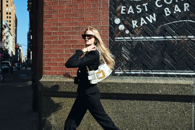 A blonde adult woman walks along a city sidewalk, wearing a black business suit and sunglasses, carrying a white purse with a gold clasp.