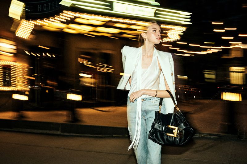 Woman in blazer and jeans poses with a purse at night in an urban environment with blurred city lights and a hotel entrance.