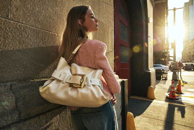 Woman in pink jacket and blue jeans poses on urban street with large white leather handbag with gold buckle, bathed in golden sunlight.