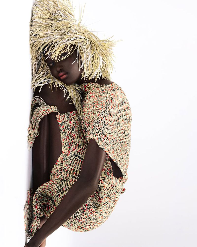 A dark-skinned model poses in a fringed, speckled dress and matching hat against a white background, showcasing high fashion.