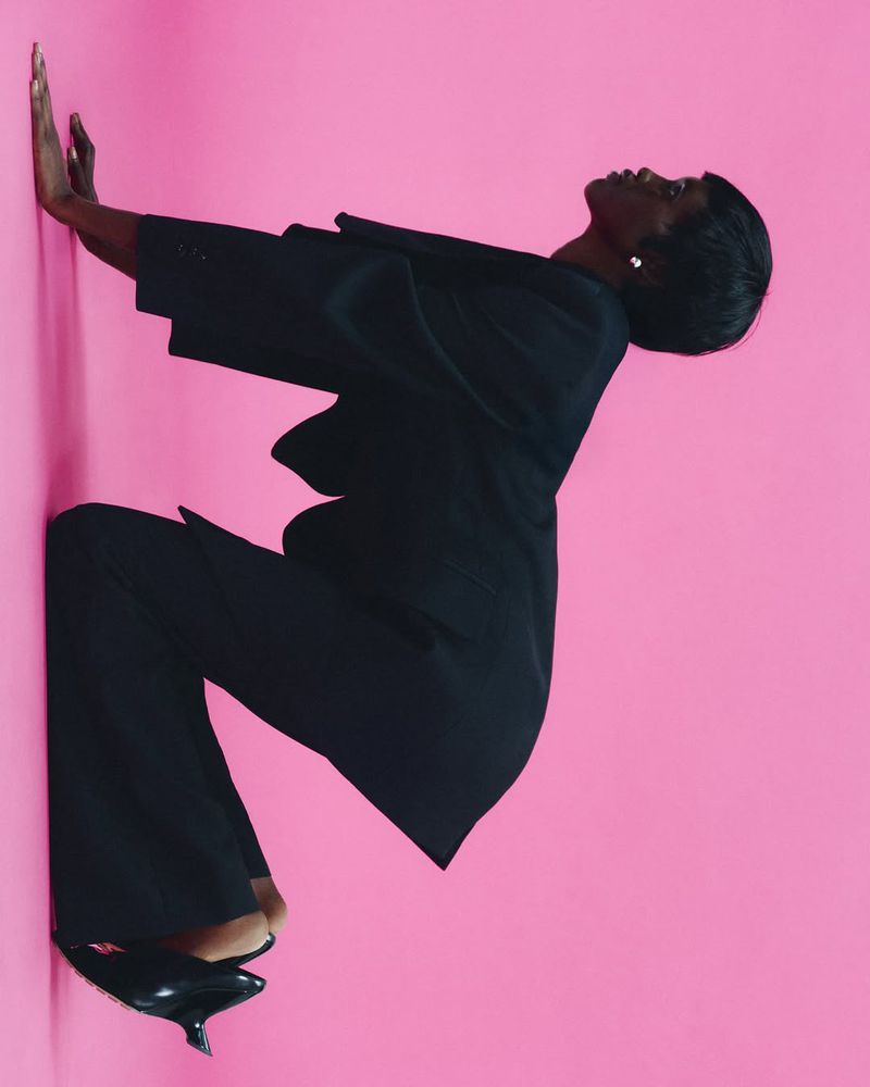 A Black woman in a black suit poses against a bright pink wall in an unusual, angled composition for a high-fashion aesthetic.