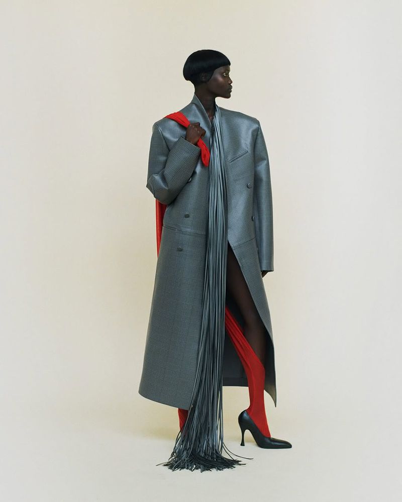 Fashionable woman in a long grey coat with fringe, red tights, and black heels against a pale backdrop in a studio setting.