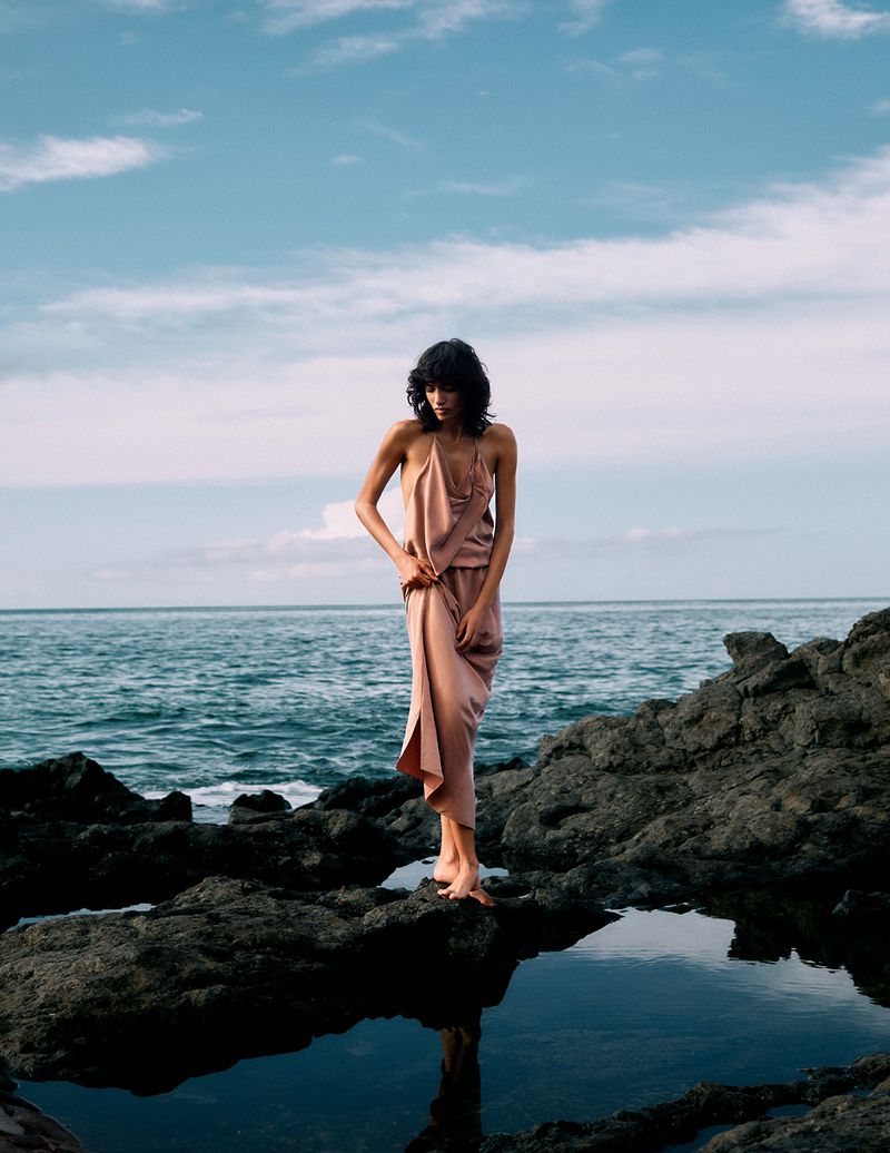A woman in a satin dress stands barefoot on a rocky beach with the ocean and a cloudy sky in the background, creating a serene fashion shot.