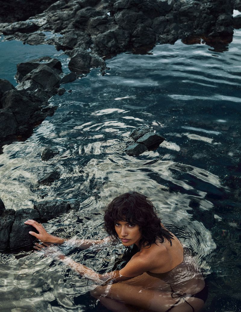 Woman sits in shallow ocean water with dark rocks. The model looks at the camera. The water is clear and the rocks are black.