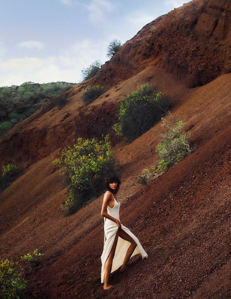 An adult woman in a light-colored dress walking on a reddish-brown hillside with green vegetation against a partly cloudy sky.