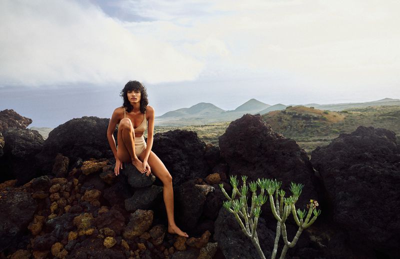 A woman in swimwear sits on dark rocks amidst a volcanic landscape with distant mountains under a cloudy sky, creating a serene outdoor scene.