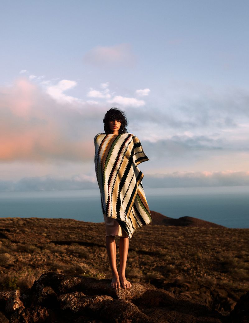 Person standing on a hill wrapped in a striped textile, with a distant ocean view and cloudy sky at dusk, contemplative mood.