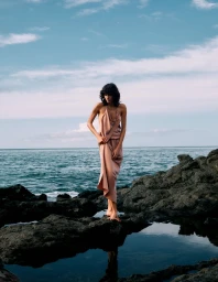 Model in a silk slip dress standing on coastal rocks by the ocean under a cloudy sky