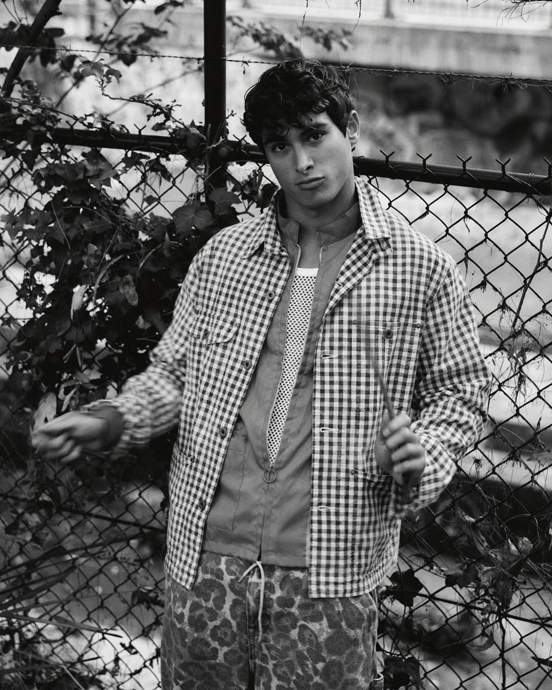 Monochrome portrait of a young adult male wearing a checkered shirt and animal-print pants standing in front of a chain-link fence.