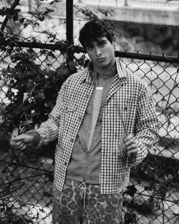 Monochrome portrait of a young adult male wearing a checkered shirt and animal-print pants standing in front of a chain-link fence.