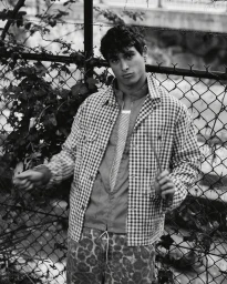 Young man standing against a chain-link fence wearing a checkered jacket and patterned trousers