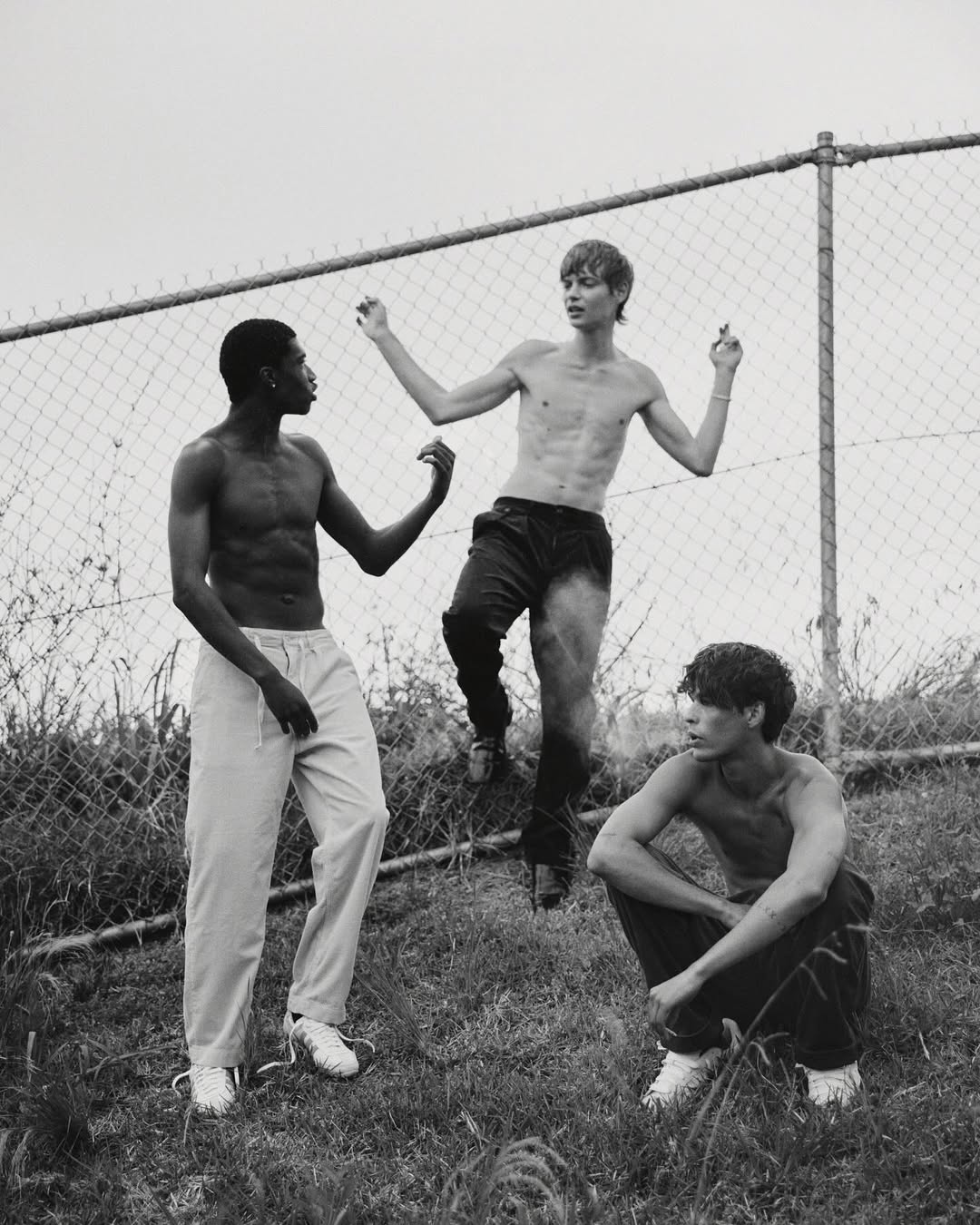 Three shirtless young men of different ethnicities stand near a chainlink fence outside, in a monochrome, high-contrast outdoor setting.