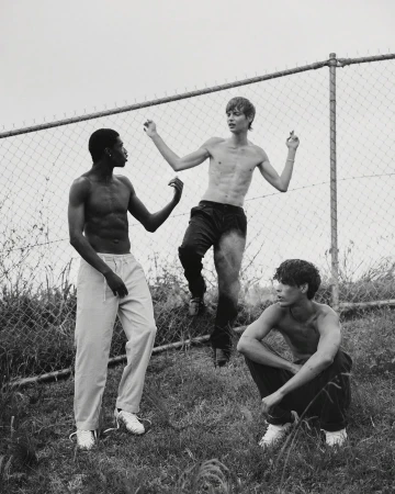 Three shirtless young men of different ethnicities stand near a chainlink fence outside, in a monochrome, high-contrast outdoor setting.