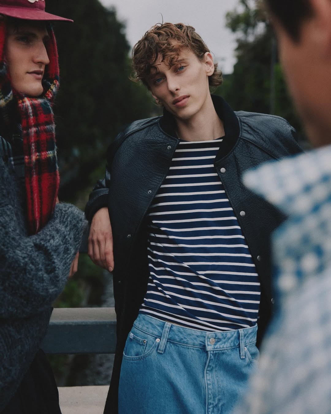 Three men in casual fashion stand outdoors with one wearing a striped shirt and jacket in a medium shot on an overcast day.