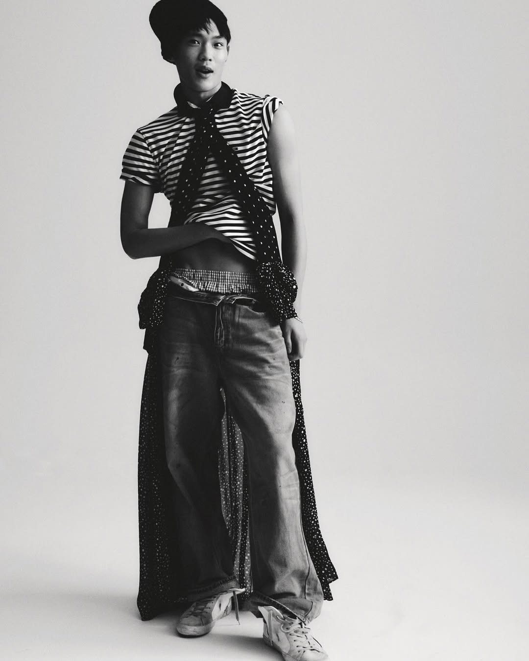 Young adult male fashion model poses in studio wearing a striped top, jeans, beanie, and polka dot scarf in a fashion editorial.
