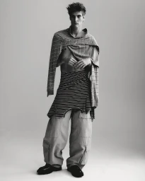 Monochrome fashion editorial portrait of a male model wearing layered striped garments and wide cargo trousers against a pale studio background.