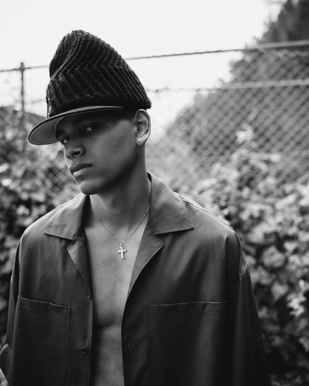 Portrait of an adult male wearing a dark cap and a shirt, standing outdoors with greenery and a fence in the background.