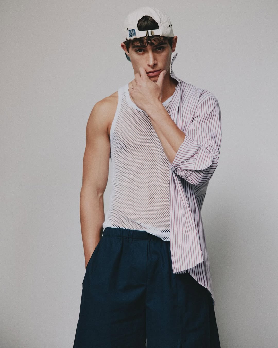 Portrait of a young adult male wearing a baseball cap, striped shirt, and mesh tank top in a photo studio with a neutral backdrop.