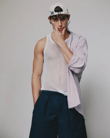 Portrait of a young adult male wearing a baseball cap, striped shirt, and mesh tank top in a photo studio with a neutral backdrop.