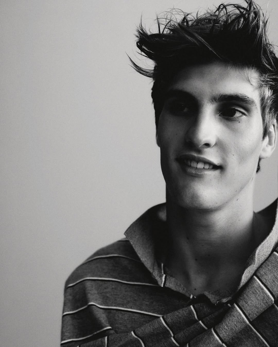 Headshot of a young adult male with dark hair in a striped shirt, against a plain light grey background.