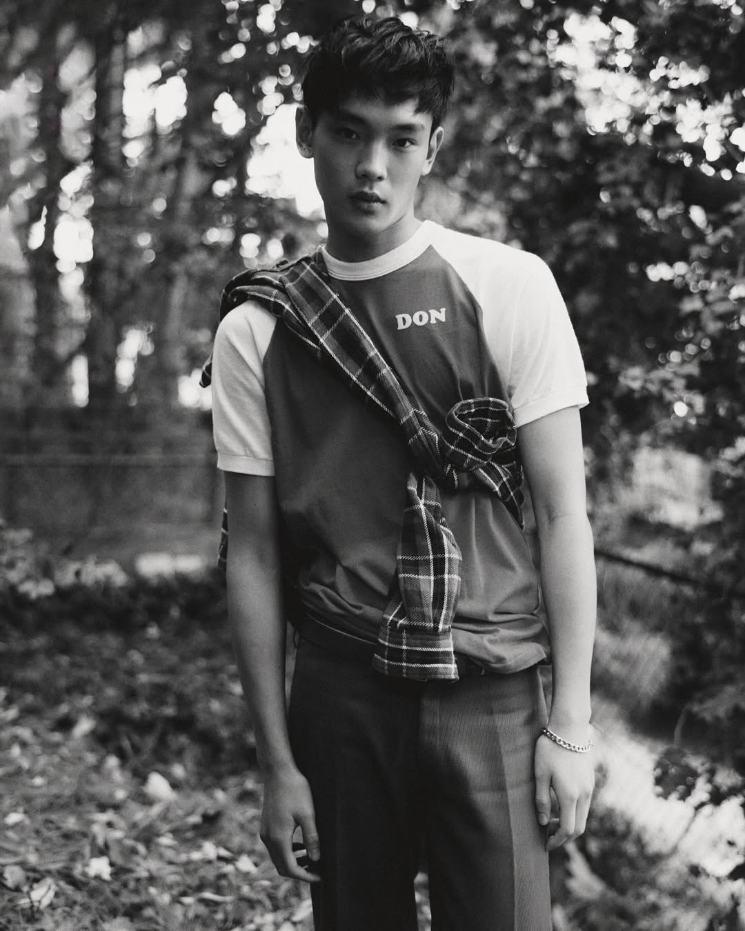 Monochrome portrait of a young adult male in a t-shirt with a plaid shirt draped over his shoulders, posing outdoors in a wooded area.