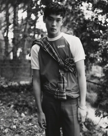 Monochrome portrait of a young adult male in a t-shirt with a plaid shirt draped over his shoulders, posing outdoors in a wooded area.