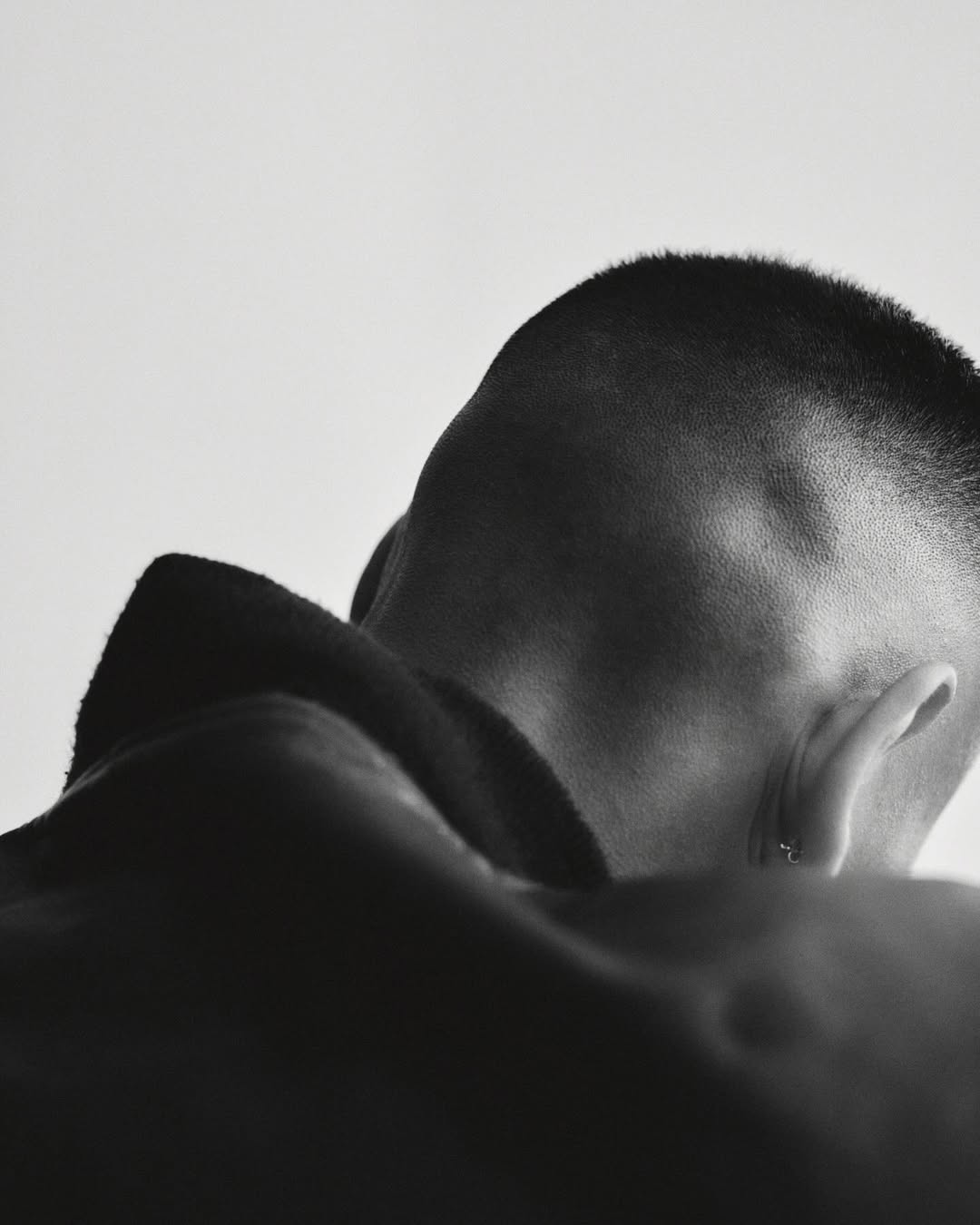 Monochromatic portrait of a bare-chested man's head and upper back featuring short hair, a silver earring, and neutral background.