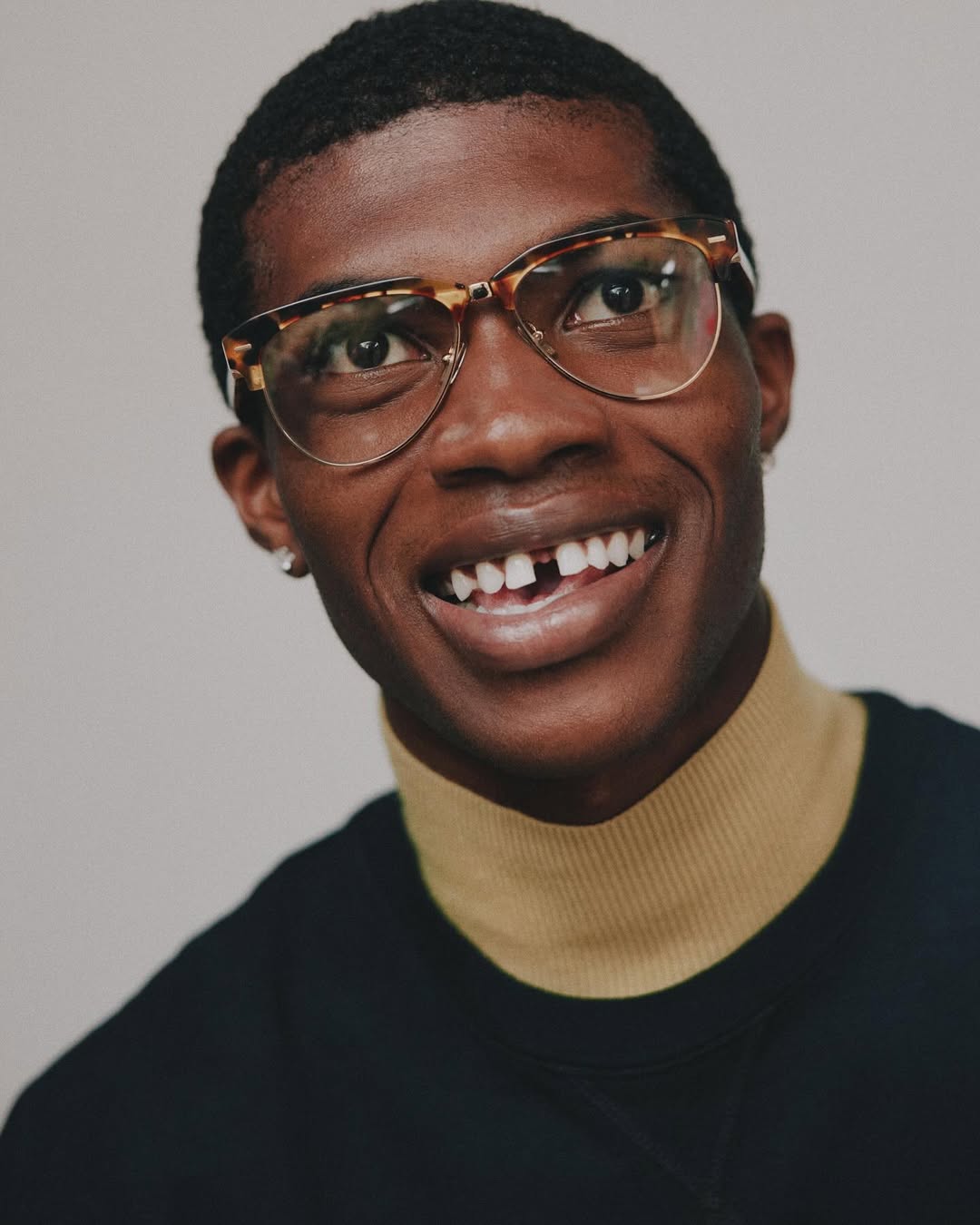 Close-up portrait of a smiling young adult male wearing glasses and a black top with a tan turtleneck, against a neutral background.