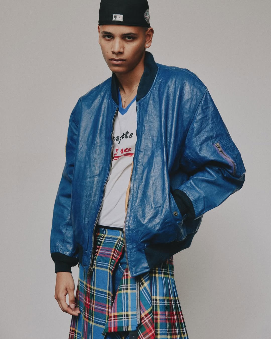 Young adult in blue leather bomber jacket and plaid kilt stands against a neutral backdrop in a fashion-forward studio portrait.
