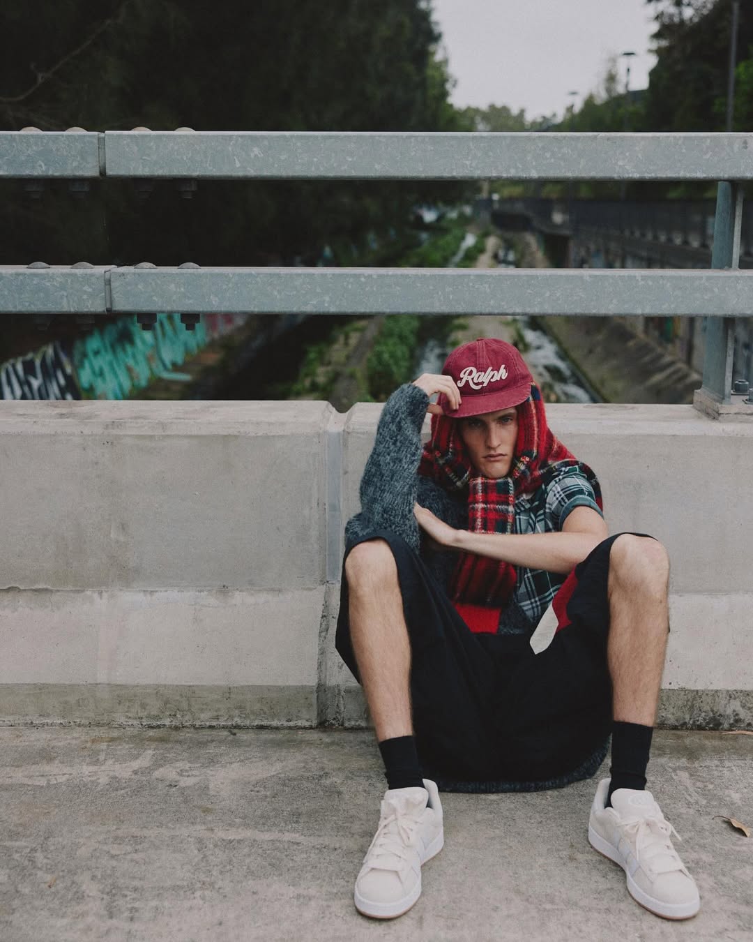 Male model in a red cap and plaid scarf sits on a concrete ledge with an urban river landscape in the background. Fashion editorial style.