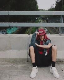 Young man wearing a red cap, plaid scarf, and shorts seated near a concrete bridge for COAT Magazine