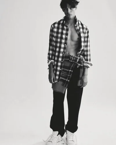 Monochrome portrait of a young man wearing an open checked shirt and trousers against a seamless plain background.