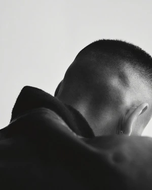 Close-up monochrome portrait focusing on the back of a man's shaved head and neck wearing a dark jacket against a plain background