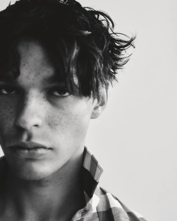 Close-up portrait of a young man with dark messy hair and freckles wearing a plaid collared shirt against a plain light background