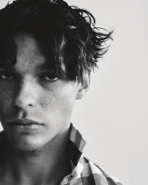 Close-up portrait of a young man with dark messy hair and freckles wearing a plaid collared shirt against a plain light background