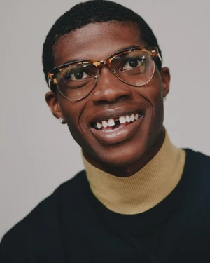 Portrait of a Black man wearing tortoise shell glasses and a dark top with a mustard turtleneck collar against a neutral grey background.