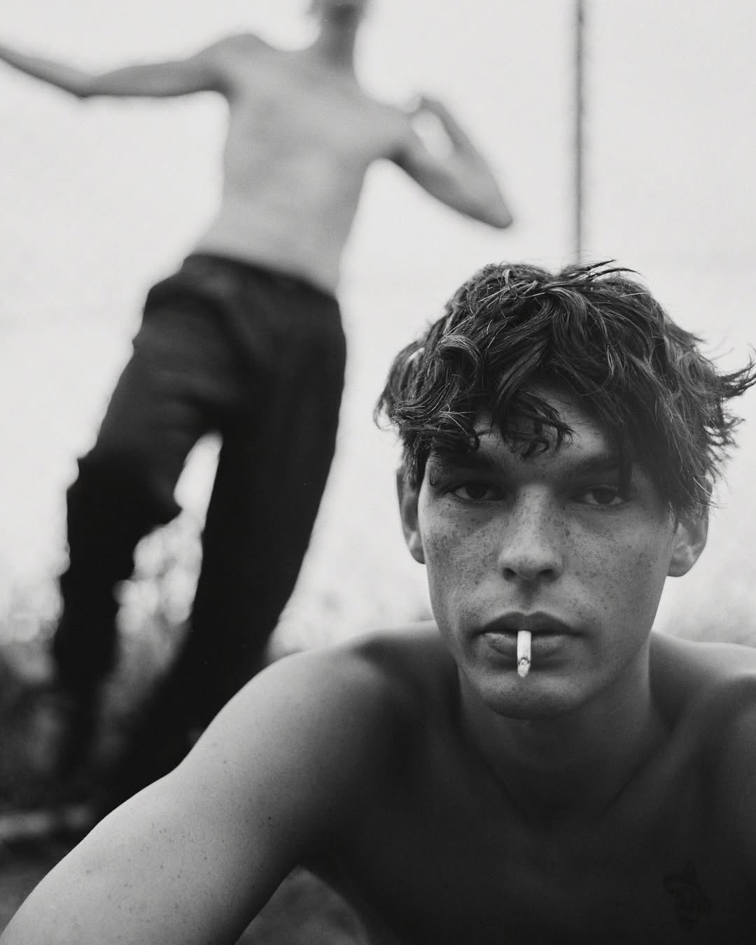 Young man with dark tousled hair and a cigarette in his mouth posing outdoors with another shirtless man blurred in background