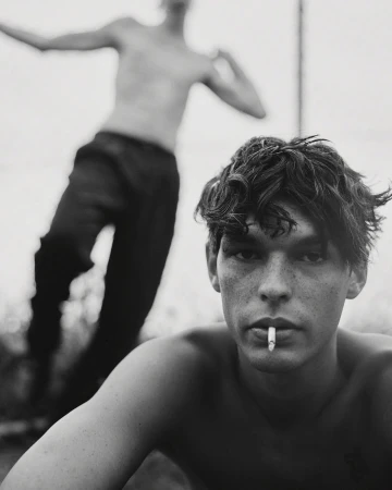 Young man with dark tousled hair and a cigarette in his mouth posing outdoors with another shirtless man blurred in background