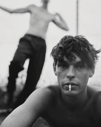 Young man with dark tousled hair and a cigarette in his mouth posing outdoors with another shirtless man blurred in background