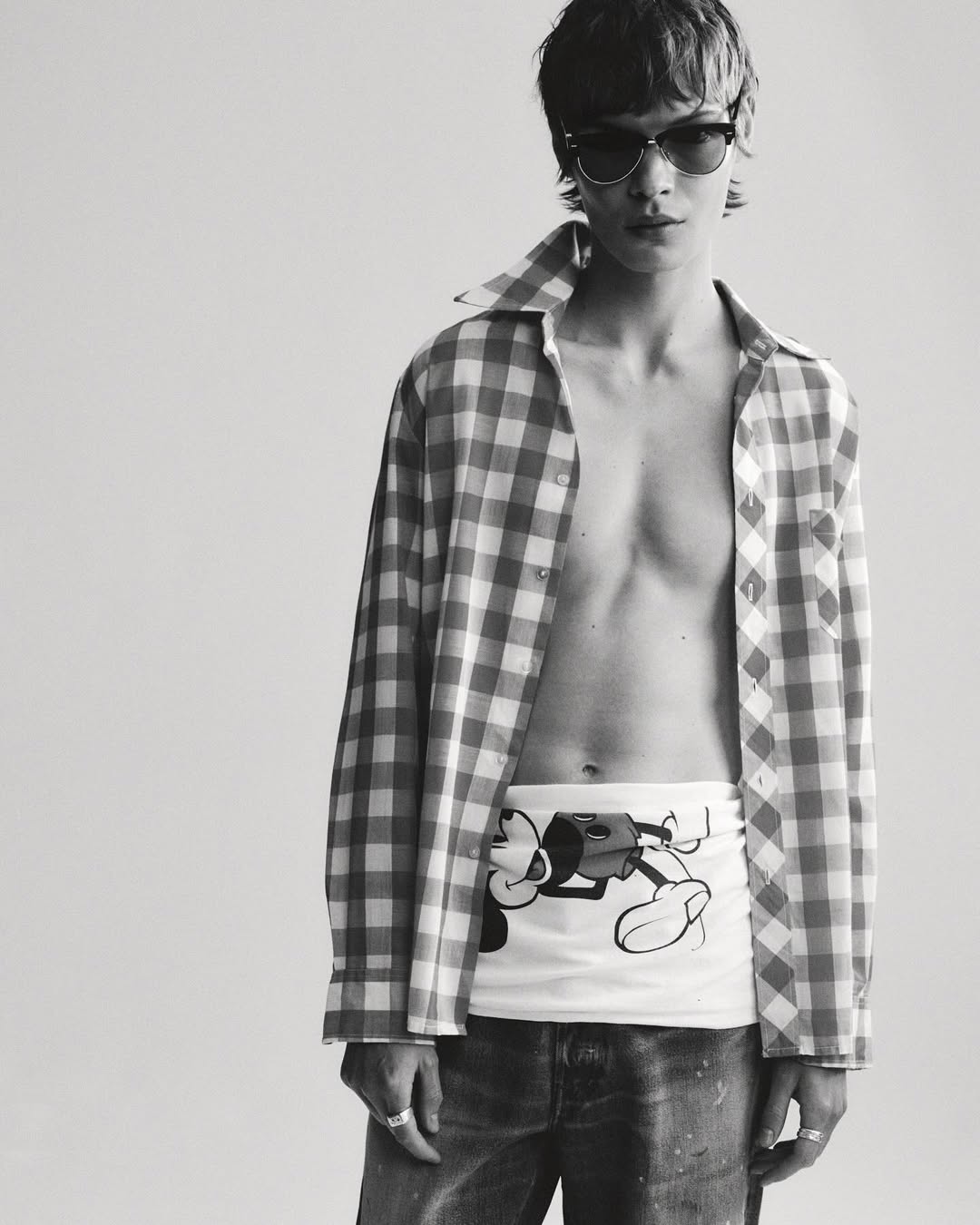 Monochrome portrait of a young adult with sunglasses, unbuttoned plaid shirt, printed top, and jeans in a bright photo studio.
