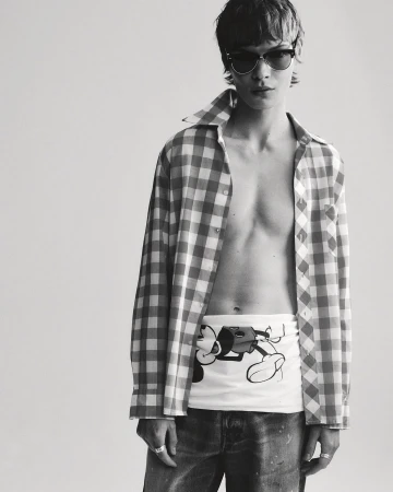 Monochrome portrait of a young adult with sunglasses, unbuttoned plaid shirt, printed top, and jeans in a bright photo studio.