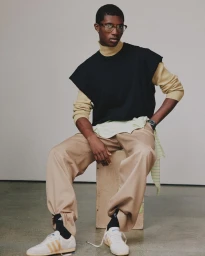 Black man wearing glasses, layered sweater vest, and tan trousers seated on a wooden crate against a neutral grey studio wall background