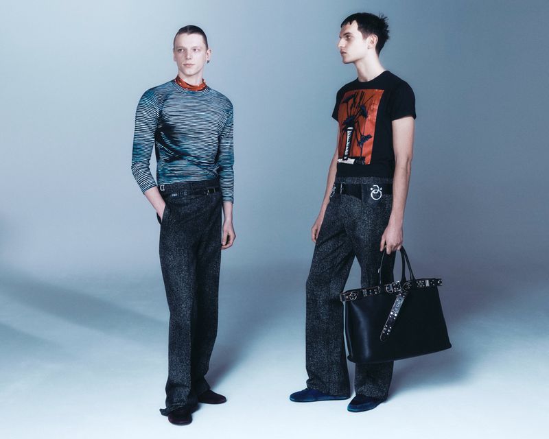 Two adult men in fashion apparel pose in a photo studio wearing graphic tees, long sleeve shirts, wide-leg trousers and carrying a leather tote.