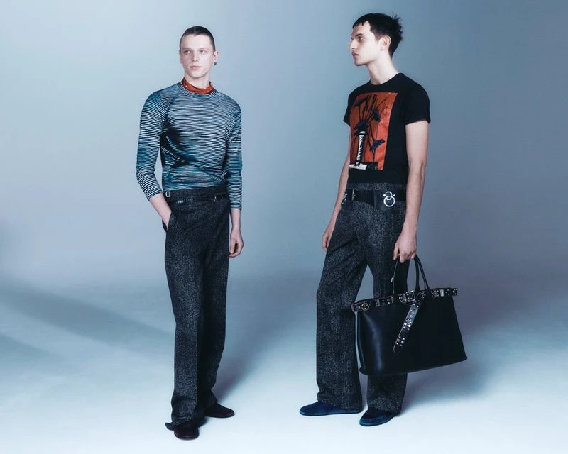 Two adult men in fashion apparel pose in a photo studio wearing graphic tees, long sleeve shirts, wide-leg trousers and carrying a leather tote.