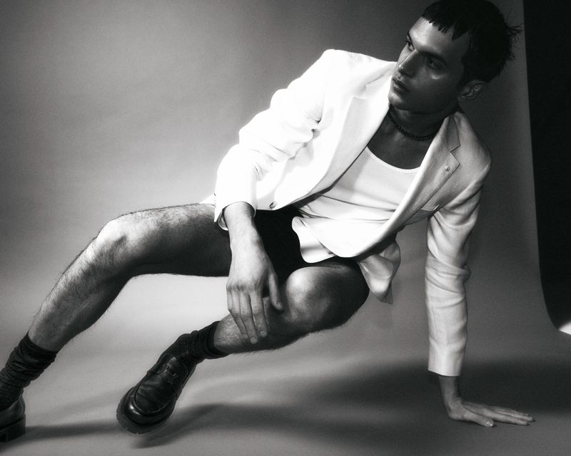 Black-and-white portrait of a person in a white blazer, black shorts, and black socks sitting on the floor looking away from the camera.
