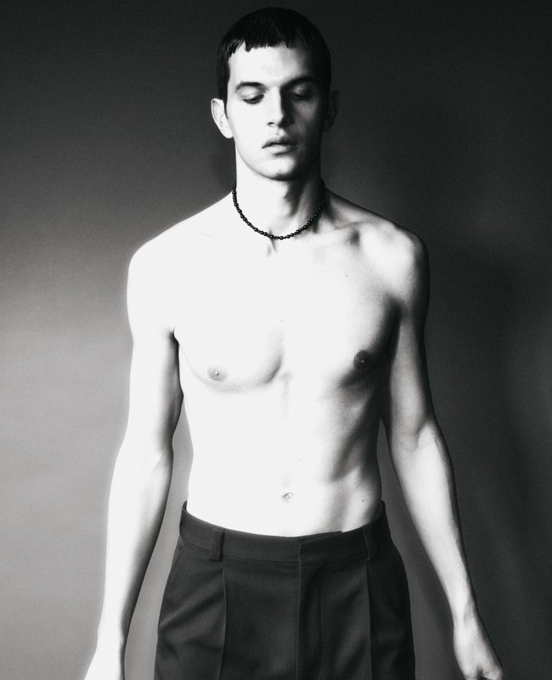 Black and white portrait of a shirtless adult male with dark hair wearing dark pants and a beaded necklace against a grey backdrop.