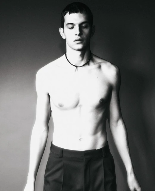 Black and white portrait of a shirtless adult male with dark hair wearing dark pants and a beaded necklace against a grey backdrop.